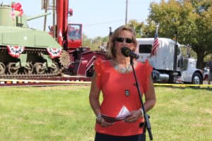 Windward Design Group - Meet Tina Marker Tina Marker speaking at outdoor event with military vehicles in background.